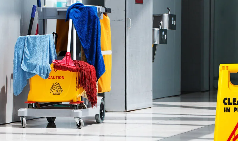 A commercial cleaning trolley with mop, towels and a wet-floor caution sign in an office corridor